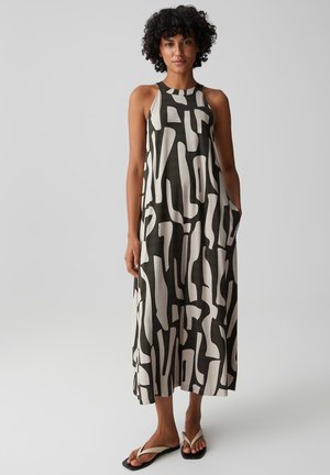 Donna con capelli ricci che indossa un vestito midi senza maniche con motivo astratto bianco e nero e infradito bianche, in piedi con le mani nelle tasche.