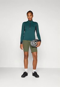 Długi rękaw rowerowej koszulki w kolorze morskim z zamkiem błyskawicznym, oliwkowozielone spodenki, czarne trampki oraz szary kask trzymany w ręce, z dopasowanym krojem.