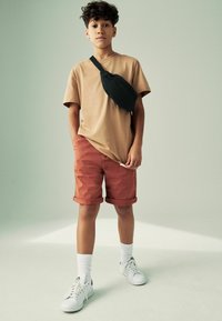 T-shirt beige, short couleur rouille, sac à bandoulière noir, baskets blanches et chaussettes blanches. Design simple avec des shorts retroussés, style décontracté.