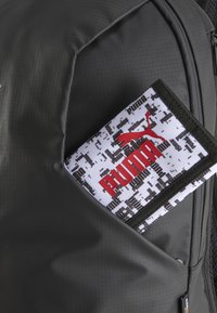 Čierny textúrovaný batoh s bielo-čiernym vzorovaným peňaženkou, na ktorej je čiastočne viditeľné červené logo Puma v vonkajšej kapsičke.