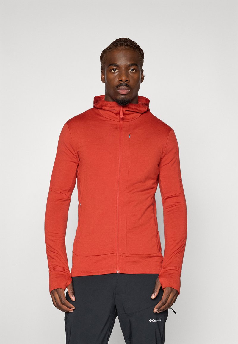 Rode zip-up hoodie gemaakt van zachte stof, met een hoge kraag, lange mouwen en duimgaten. Gecombineerd met donkere broek voor contrast.