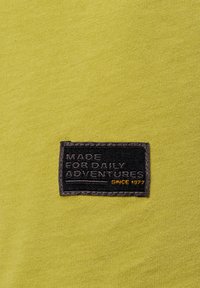 Olijfgroen stof met een gladde textuur, voorzien van een zwart etiket met reliëftekst: "MADE FOR DAILY ADVENTURES" en "SINCE 1977."