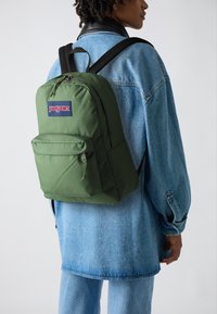 Zöld Jansport hátizsák, elülső zsebbel és húzózsinórral, világoskék farmerdzsekivel viselve. Az anyag sima, struktúrált formával rendelkezik.