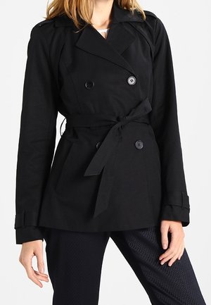 Femme portant un trench-coat noir croisé avec une ceinture nouée à la taille et un pantalon noir texturé, debout avec une main détendue.