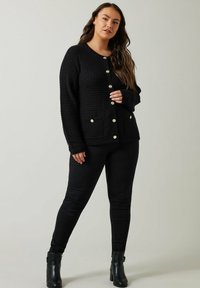 Zwarte gebreide cardigan met een textuurpatroon, voorzien van gouden knopen en twee voorzakken, gecombineerd met zwarte broek en enkellaarsjes.