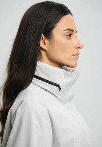 Chaqueta gris claro con un cuello alto, que presenta un bolsillo con cremallera negra en el costado. Textura suave, diseño simple y un corte relajado.
