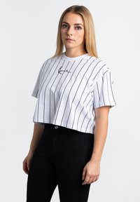 Abgeschnittenes weißes T-Shirt mit schwarzen vertikalen Streifen und kleinem gesticktem Logo auf der Brust. Kurze Ärmel und runder Ausschnitt.