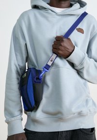 Borsa a tracolla blu con materiale a rete e solido. Tracolla regolabile con fibbia, dettaglio di piccola tasca, indossata sopra una felpa con cappuccio grigio chiaro.