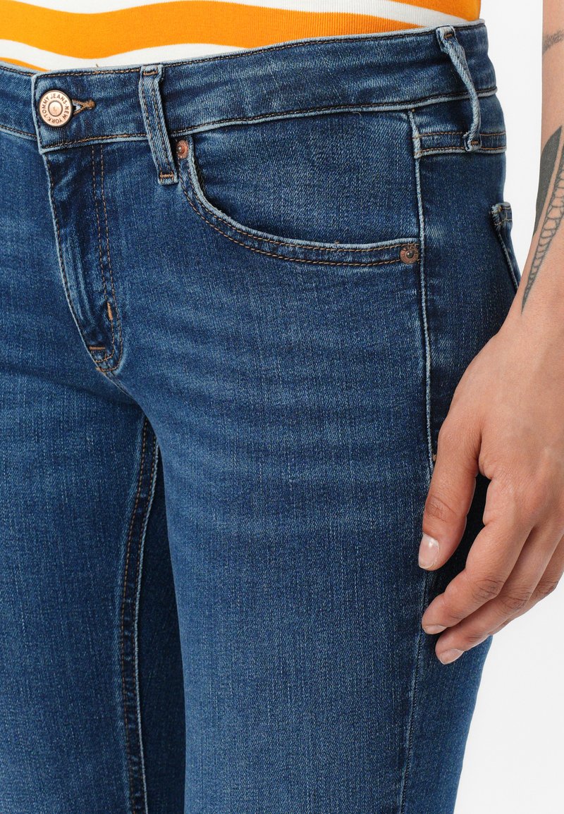 Fasowane niebieskie dżinsy z średnim stanem, pięcioma kieszeniami i pomarańczowymi szwami wzdłuż szwów. Zwężany krój nogawki.