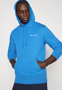 Μπλε βαμβακερό φούτερ με κουκούλα και εμπρός τσέπη, ρυθμιζόμενο κορδόνι στη κουκούλα και κεντημένο λογότυπο "Champion" στο στήθος. Ριμπ μανσέτες και τελείωμα.
