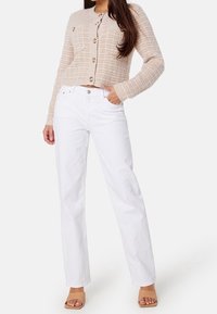 Cardigan beige texturé avec des boutons dorés, associé à un jean droit blanc. Le modèle porte des sandales à talons blocs nude, les mains dans les poches.