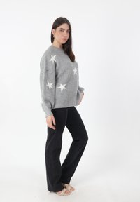 Maglione grigio lavorato a maglia con motivi a stelle bianchi, scollatura tonda e spalle scese, abbinato a jeans neri e sandali chiari.