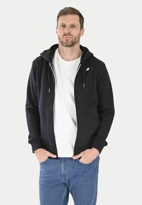 Haut à capuche zippé noir avec un logo blanc sur le côté gauche de la poitrine, présentant une capuche avec cordon, des poignets côtelés et une texture lisse.