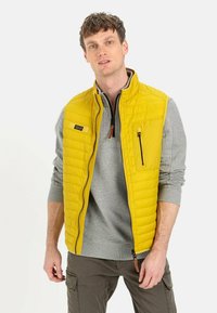 Gul quiltet vest med lynlås, der har en frontlomme og en grå langærmet skjorte under. Tekstureret stof og farvekontrast er til stede.