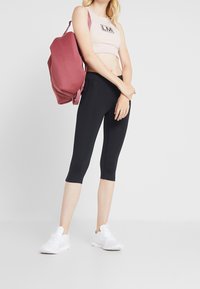 Lichtroze crop top met zwart logo, zwarte capri legging, en witte sneakers, aangevuld met een gestructureerde bordeauxrode rugzak.