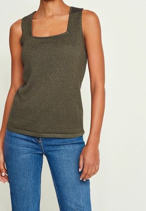 Femme portant un top en tricot sans manches vert olive à encolure carrée et un jean en denim bleu, debout devant un fond clair uni.
