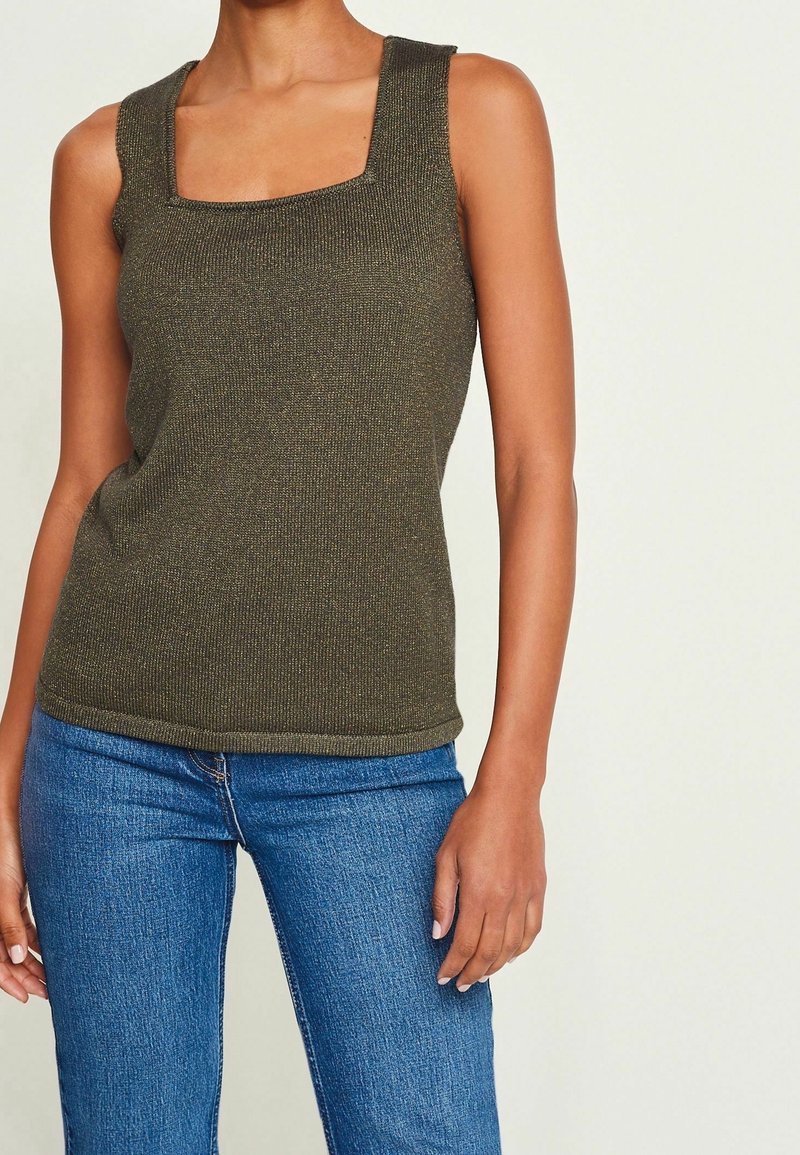 Femme portant un top en tricot sans manches vert olive à encolure carrée et un jean en denim bleu, debout devant un fond clair uni.