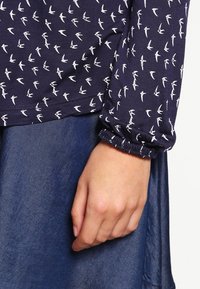 Tummansininen pitkähiha shirtti, jossa on valkoisia lintukuviota, rypistetyt hihansuut, yhdistettynä farkkukankaisiin alaosiin. Pehmeä tekstuuri, rento muotoilu.