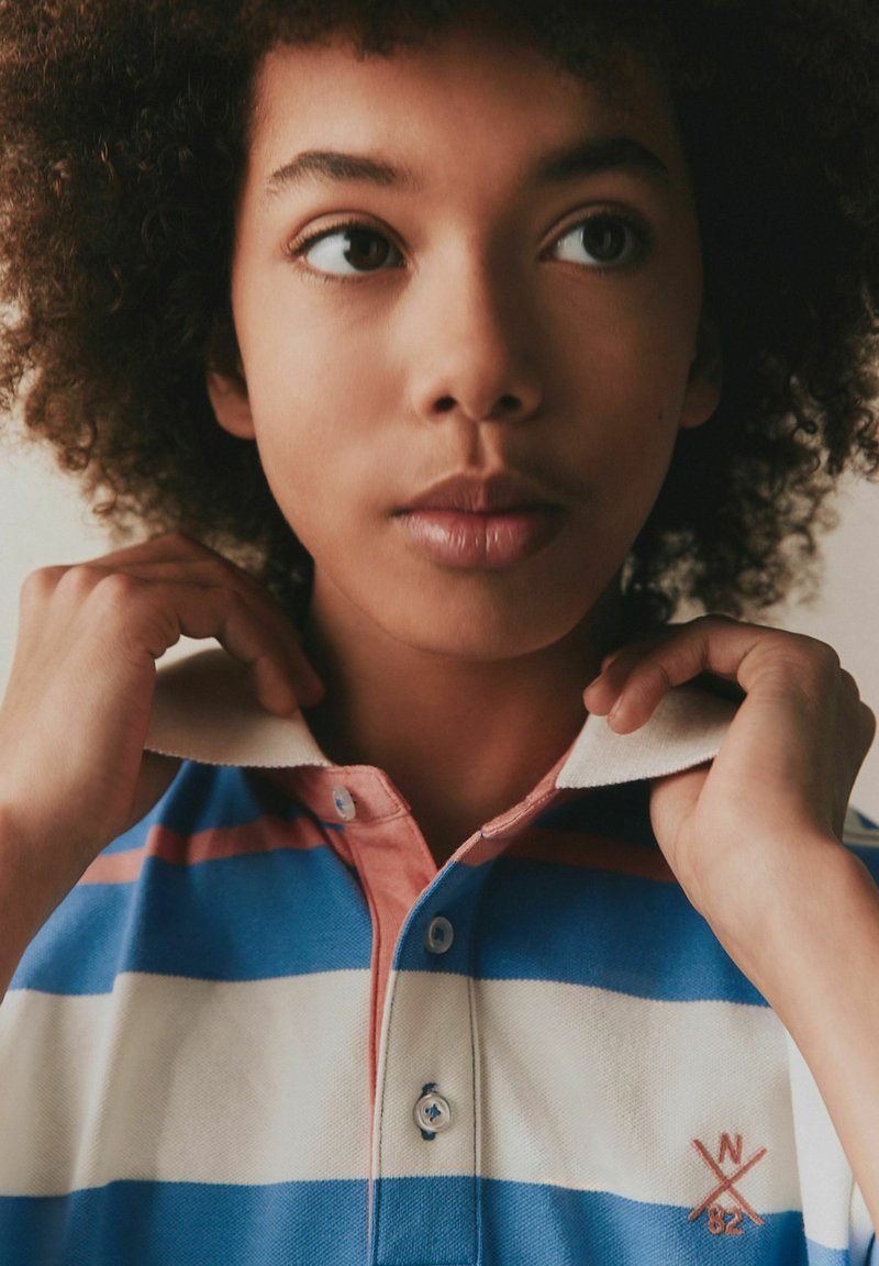 Młoda osoba poprawiająca kołnierzyk koszulki polo w paski w kolorach niebieskim, białym i różowym, z wyszywanym napisem "W 82" na piersi.