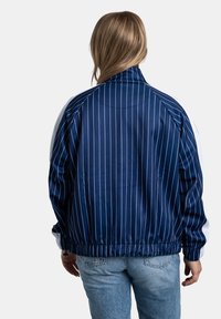 Granatowa kurtka z białymi paskami, elastycznymi mankietami i talią. Gładki, błyszczący materiał; zawiera prążkowany kołnierz i kontrastowe białe akcenty.