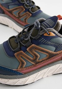 Baskets de sport bleues et vertes avec un dessus en synthétique texturé, des accents marron contrastants, des lacets réfléchissants et une semelle coussinée prononcée.