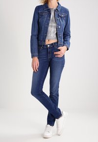 Džínová bunda s přiléhavým střihem, tmavě modré barvy, se dvěma náprsními kapsami, spárovaná s vysokými tmavými džíny a bílými teniskami.