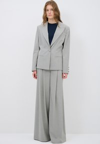 Lichtgrijs op maat gemaakt pak met een blazer met één knoop, wijde broek en een donkerblauwe top. De stof heeft een subtiele textuur en drapering.