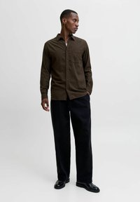 Man wearing brown button-up shirt, black wide-leg pants, and black dress shoes standing with one hand in pocket, looking right.