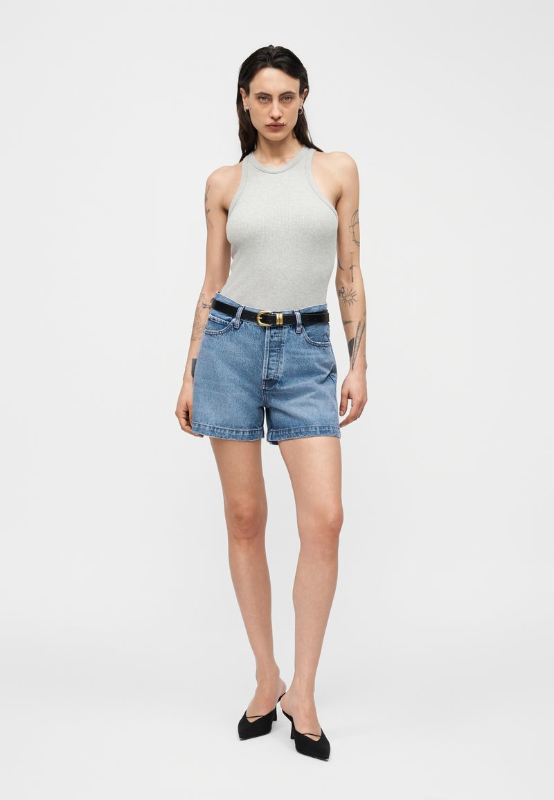 Frau steht, trägt graues ärmelloses Oberteil, Jeansshorts mit schwarzem Gürtel und schwarze Absatzschuhe, Tätowierungen an den Armen sichtbar.