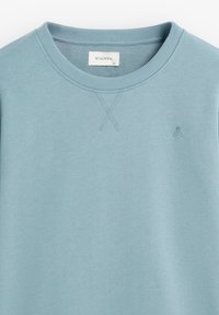 Sudadera azul claro con cuello redondo y puños acanalados, que presenta un pequeño logo en el pecho y detalle de pespunte en el escote.