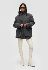 Donna che indossa un blazer oversize grigio scuro, una gonna abbinata, stivali bianchi alti fino alla coscia, grandi orecchini d'argento, occhiali da sole scuri e tiene tra le mani i bottoni del blazer.