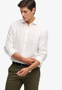 Joven ajustándose el puño de una camisa blanca abotonada, vistiendo pantalones oliva con cinturón trenzado negro y reloj de cuero marrón.