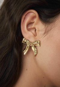 Goldfarbene Ohrringe in Form einer Schleife, mit einem strukturierten Design und fließenden Ribbons, die am Ohrläppchen eines Modells befestigt sind.