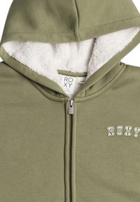 Olívovo zelená bunda s kapucňou vyrobená z mäkkej tkaniny, s predným zipsom a plyšovým bielym podšívkou. Logo "Roxy" je vyšívané na hrudi.