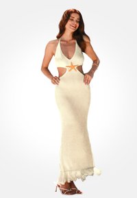 Mujer sonriente de pie con vestido de punto color crema tipo halter con cortes laterales, detalle de estrella de mar en la cintura, bajo con flecos y tacones blancos con tiras.