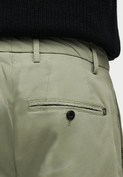 Κοντινό πλάνο ελαιδί παντελονιών που δείχνει πίσω τσέπη με κουμπί και λεπτομέρεια φερμουάρ, φορεμένα με μαύρο πλεκτό τοπ.