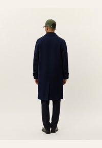 Navy wool coat with a straight cut, long sleeves, and buttoned cuffs. Worn over dark trousers and a green cap. Simple, clean design.