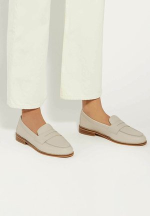 Beige leather loafers with a stitched apron design and a low wooden heel. Paired with wide white trousers, showing a clean, modern look.