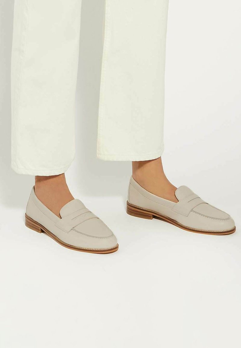 Beige leather loafers with a stitched apron design and a low wooden heel. Paired with wide white trousers, showing a clean, modern look.