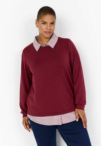 Jersey burdeos con mangas largas y escote redondo; presenta un cuello y un dobladillo a rayas en contraste, superpuesto sobre una camisa blanca a rayas.