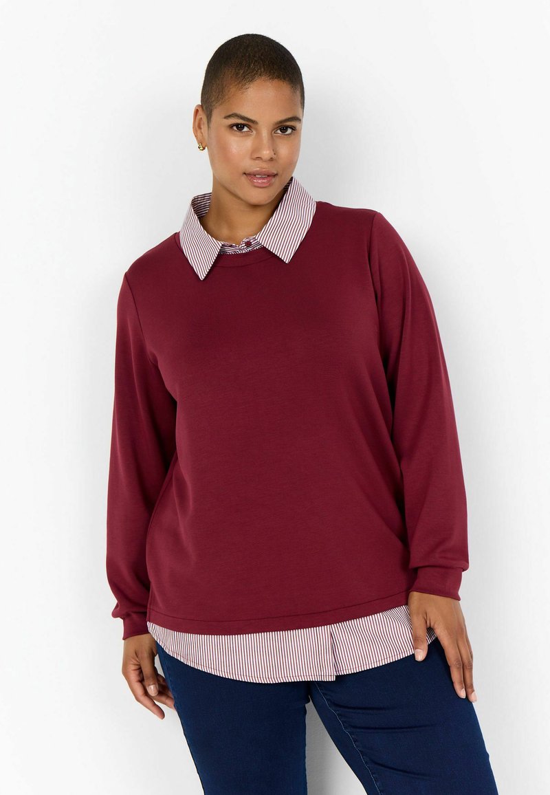 Pullover bordeaux con maniche lunghe e scollatura rotonda; presenta un colletto e un orlo a strisce in contrasto, indossato sopra una camicia a righe bianche.
