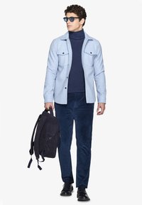 Lysblå skjortejakke over en mørk turtleneck, marineblå kordfløjl bukser og sorte sko. Holder en mørk ruskinds rygsæk.