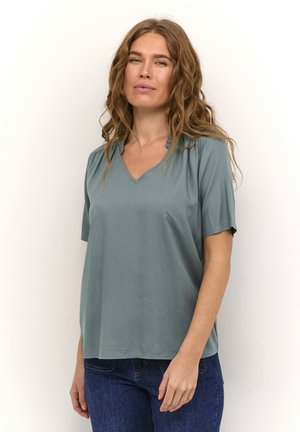 Camiseta de manga corta de pico en color azul verdoso, con un corte relajado y detalle de hombro fruncido, fabricada en un tejido suave. Combinada con jeans de mezclilla azul.