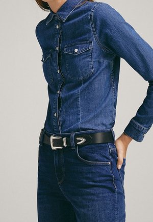 Person wearing a fitted dark blue denim shirt with buttoned breast pockets and matching dark blue denim jeans with a black belt.