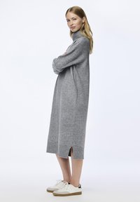 Γκρι, πλεκτό φόρεμα με ψηλό λαιμό και πλάγιες σχισμές, μεσαίου μήκους. Συνδυάζεται με λευκά αθλητικά παπούτσια με σκούρες λεπτομέρειες. Μαλακή υφή, χαλαρή εφαρμογή.