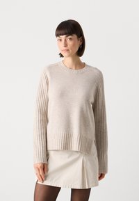 Beige strikkegenser med ribbestrikkede ermer og kant, rund hals og sidespalter. Brukt over et lysfarget skjørt.