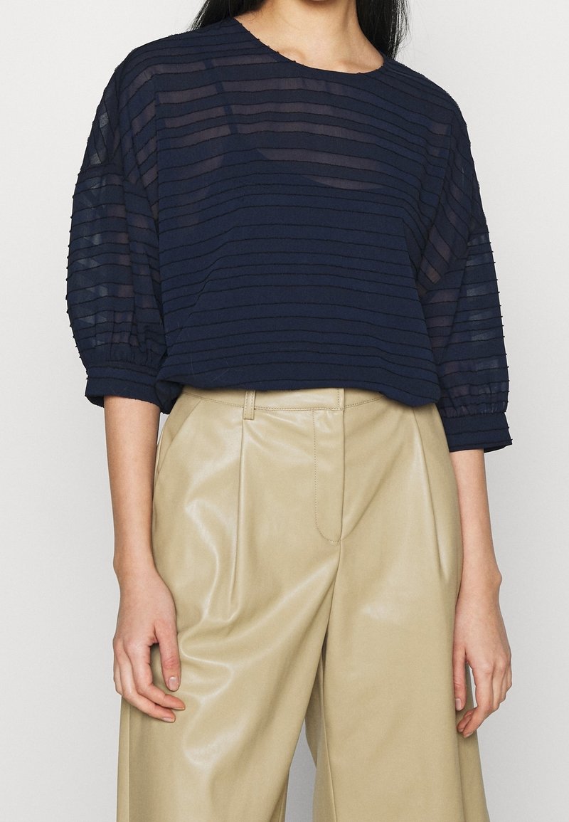 Haut rayé bleu marine, coupe ample avec manches trois-quarts ; associé à un pantalon beige plissé taille haute en similicuir.