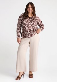 Blusa a maniche lunghe con un motivo astratto marrone e crema, abbinata a pantaloni beige a gamba larga e sandali con tacco marroni.