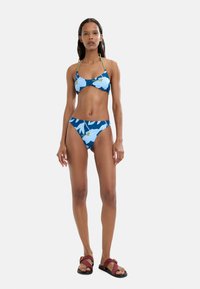 Niebieski kostium kąpielowy w kwiaty z jasnoniebieskimi kwiatami, regulowane ramiączka i pasujące majtki z wysokim stanem, zestawione z bordowymi sandałami z klamrą.