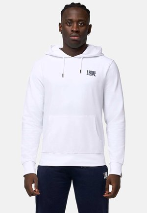 Homme portant un sweat à capuche blanc avec le logo "LEONE 1947" et un pantalon foncé avec un logo assorti, debout devant un fond uni.